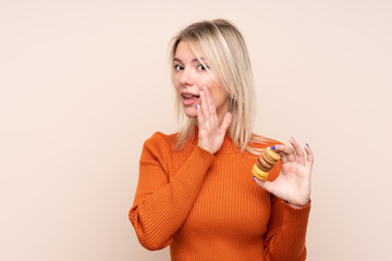 Young blonde Russian woman over isolated background holding colorful French macarons and whispering something