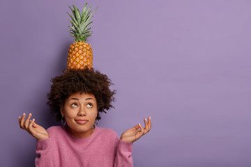 Horizontal view of black curly woman tries to maintain equilibrium, holds fresh pineapple on head, spreads palms, looks above, stands with exotic fruit against purple wall, copy space area aside - Powered by Adobe