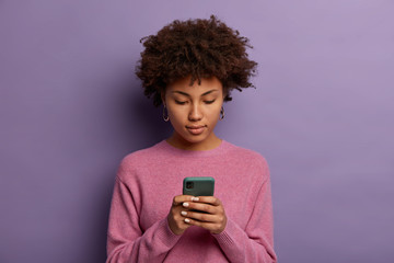 Serious looking Afro American woman reads message on modern mobile phone, surfes social media, has concentrated look in display, wears casual jumper, isolated over purple wall, updates information