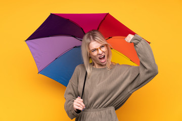 Young blonde woman holding an umbrella over isolated yellow wall celebrating a victory