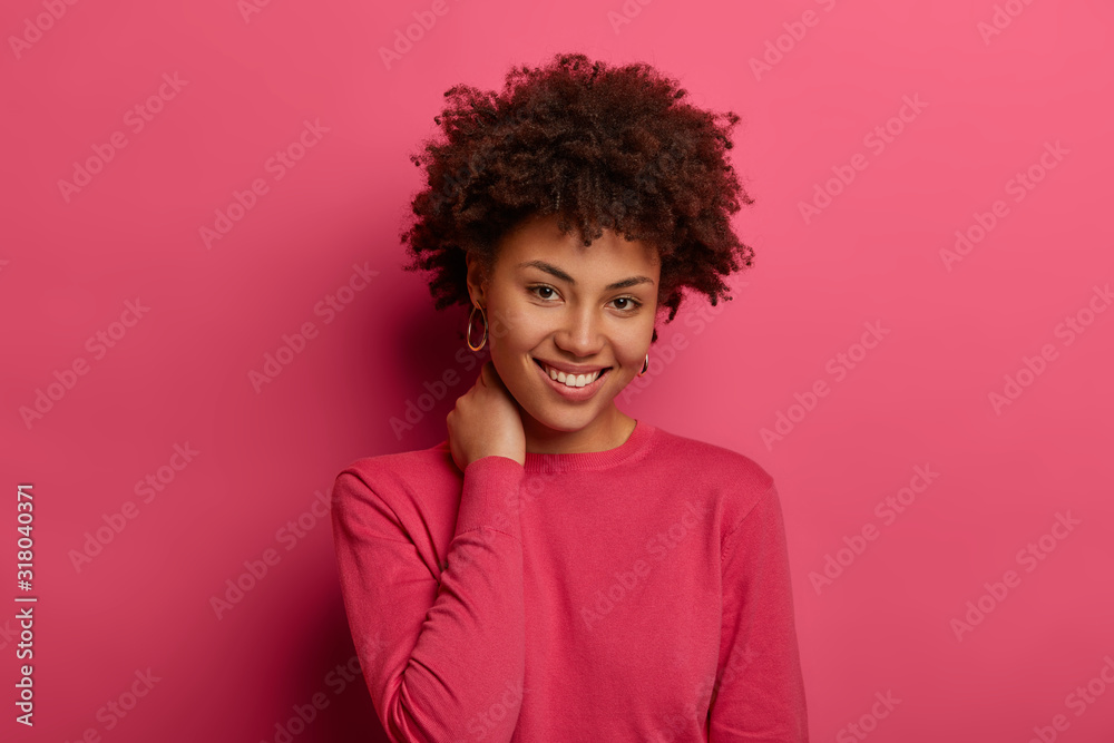Wall mural Good looking woman with curly hair, keeps hand on neck, smiles gently, wears crimson casual jumper, looks gladfully at camera, isolated over pink studio wall, has optimistic view. Happiness and joy - Wall murals