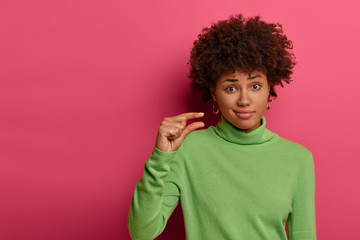 Fototapeta na wymiar Not much will be enough. Unimpressed curly woman shapes small tiny object, needs little more help and attention, wears green neck sweater, gestures indoor against pink background. Size concept