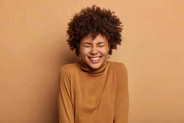 Fototapeta na wymiar Portrait of joyous glad lady with toothy smile, laughs out as hears comic story, being in high spirit, enjoys happy day, has natural curly hair, isolated over beige background. Upbeat emotions