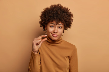 Fototapeta na wymiar Portrait of attractive woman with curly Afro hair, shapes tiny and small thing, talks about size, dressed in casual brown jumper, being unimpressed, isolated over beige studio wall. Little object