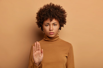Fototapeta na wymiar Stop right here. Serious dark skinned woman stands with outstretched hand, makes prohibition gesture, forbids something, smirks face, wears casual brown neck sweater, isolated on beige wall.