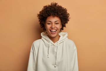 Optimistic teenage girl dressed in casual white sweatshirt, smiles happily, stands against brown background, hears funny joke from friend, gazes friendly at camera, stands indoor. Studio portrait