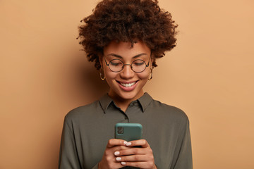 Headshot of curly woman wears transparent glasses, grey shirt, uses free online app on smartphone, views images, wears round glasses and shirt, isolated over beige background, browses social media