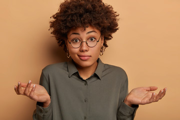 Careless hesitant woman shrugs shoulders, spreads arms sideways, cant help or has doubts, wears stylish shirt and big glasses, unable to give answer on confusing question, models in brown studio