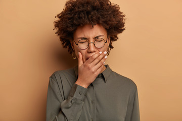 Sleepy curly woman yawns with closed eyes, covers mouth, being tired, has lack of sleep and energy, needs rest, suffers from insomnia, wears stylish shirt, poses against brown studio background