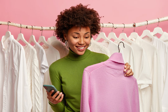 Positive black woman chooses sweater to buy, holds hanger with purple turtleneck, mobile phone in other hand, stands against white clothes hanging in wardrobe closet, has happy smile on face