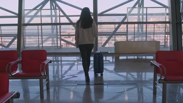 A woman with a suitcase at the airport goes to the window and looks out.