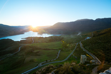 Star sun at dawn in a green mountain landscape, with a lake.