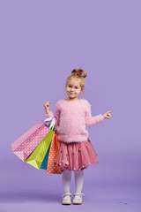 Portrait of a happy little child girl standing on a purple background holding shopping bags, package. Looking at the camera.