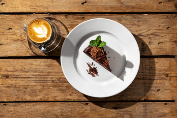 Overhead view of piece of chocolate cake with cappuccino on wooden table under harsh sunlight