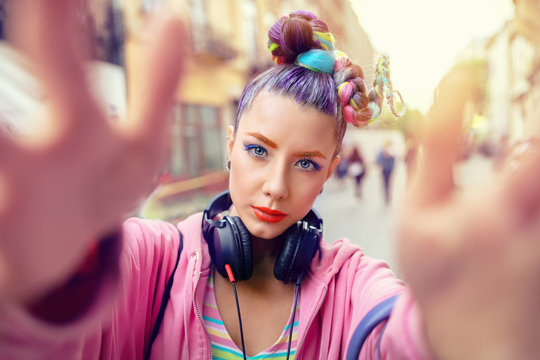 Playful cool rebel funky hipster young girl with headphones and crazy hair taking selfie on street