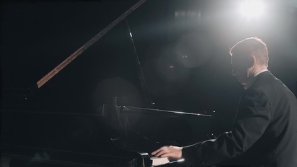 Pianist in black suit masterfully playing the grand piano on big stage in concert hall with dimmed light