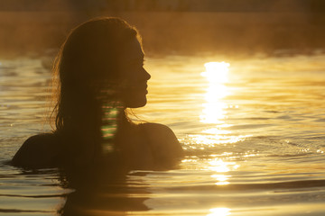 Fototapeta premium silhouette of a beautiful girl with wet hair in water, head of a woman bathing in a hot spring at sunrise, summer relaxation and vacation concept