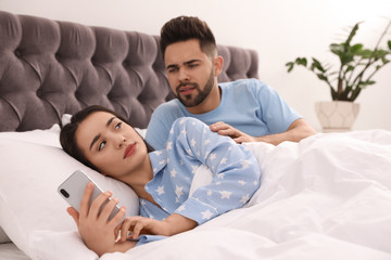 Fototapeta premium Young woman ignoring her distrustful boyfriend while using smartphone in bed at home