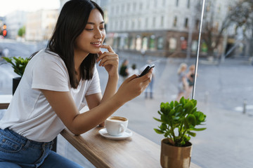 Fototapeta premium Leisure, weekends and urban lifestyle concept. Carefree young modern girl using smartphone, scroll social media as sitting alone cafe, lean coffee table, drink cappucino smiling, look mobile display