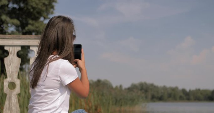 Beautiful girl in hipster outfit sits on the boards of wooden pier on the shore of a lake or river. Young woman uses her phone. She takes pictures and sends messages over the Internet via smartphone.