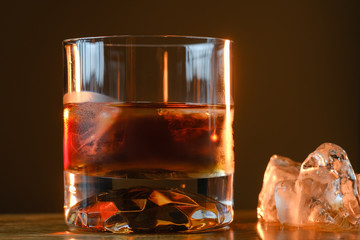 Glass of whiskey with ice cubes served on wooden board. Dark background, glossy golden color hard liquor on countertop with highlight. Night life bar relaxation concept