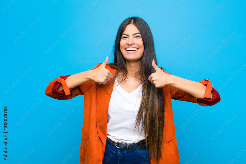 Wall mural young pretty latin woman smiling broadly looking happy, positive, confident and successful, with both thumbs up against flat wall