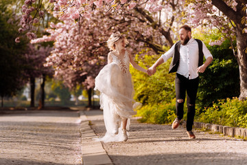 Fototapeta premium Happy stylish man with beard and woman with long dress are having fun in spring blossoming sakura park. Newly wedded hipsrers couple in the park. Just married. run in the park and hold hands.