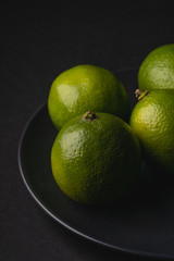 Lime sour fruits in black plate on moody dark background, angle view close up, vitamins and healthy food