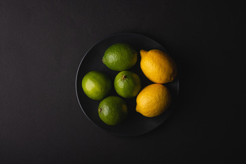 Lime and lemon sour fruits in black plate on moody dark background, top view, vitamins and healthy food