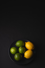 Lime and lemon sour fruits in black plate on moody dark background, angle view, vitamins and healthy food