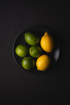Lime and lemon sour fruits in black plate on moody dark background, top view, vitamins and healthy food