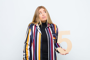 young pretty blonde woman confused, doubtful, thinking, holding a number 5.