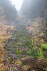 Scenic mountain forest waterfall, red rocky cliff covered by sunlit green moss and thick morning mist