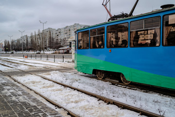 Naklejka na ściany i meble Modern tram riding in winter city. Contemporary blue and green tram riding on snowy rails on cloudy winter day in city