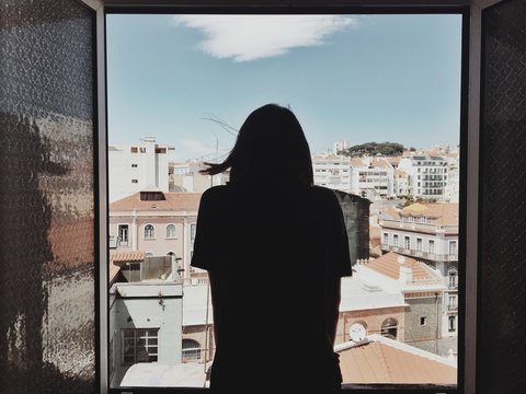 Rear View Of Woman Looking Through Window By Buildings Against Sky