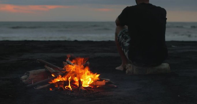 A young guy gets up from the fireplace and leaves the fire. Young urban man sit near the fire in the evening. Handsome hipster male at sunset near the bonfire on the empty sand tropical beach.