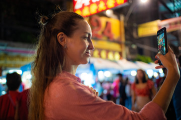 Young caucasian tourist women take photo smartphone travel in street night market blurred people background