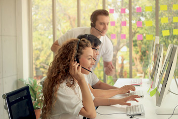 Young beautiful woman with headphones working at call center service desk consultant with her teammates while team supervisor helping new teammate and training junior to talk with the customer on hand