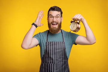 Portrait of amazed young chef man holding alarm clock and gesturing over yellow background