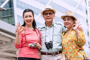 Group elderly traveler with binoculars and camera showing okay gesture while standing together in city. Senior couple with sister happiness in trip on weekend. Lifestyle retirement and family concept.