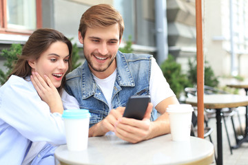 Attractive young couple in love drinking coffee while sitting at the cafe table outdoors, using mobile phone.