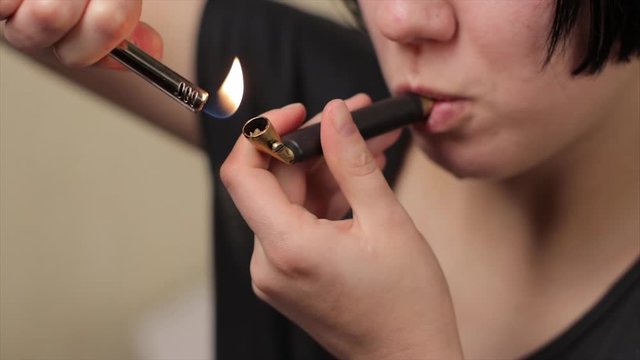 Young girl or teen lighting and smoking brass pipe with medical marijuana weed indoors and exhaling smoke in slow motion