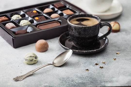 Box of Luxury Chocolate candies selection with cup of black coffee and silver spoon on light table background.