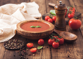 Wooden plate of creamy tomato soup with wooden spoon, pepper and kitchen cloth on wooden background with raw tomatoes and basil.