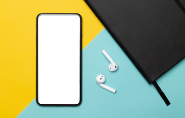 Black smartphone with blank screen and wireless earphones and notebook isolated on yellow and blue background. Clipping path.