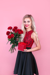 Blonde girl posing and smiling on bright pink background. Lady with red flowers and gift box in her hands
