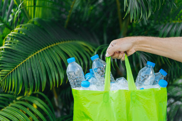 Bag with plastic bottles in a man's hand against the background of tropical summer greenery. Plastic collection, environmental protection.