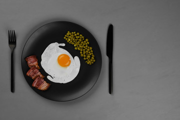 English breakfast fried eggs, bacon,  green pea on black plate, grey background. black fork and knife. top view