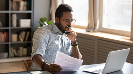 Focused african businessman holding documents looking at laptop doing research