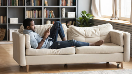 Obraz premium Serious relaxed millennial african man lying on couch using smartphone